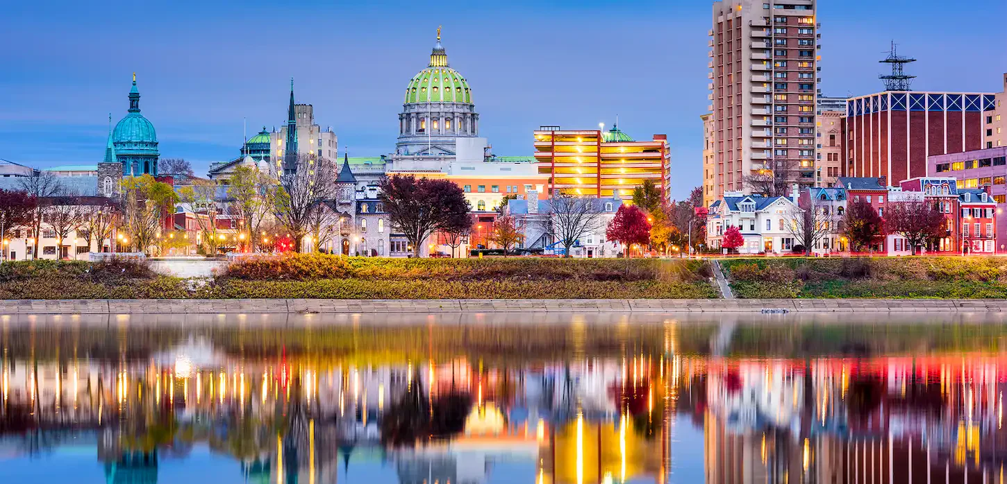 Harrisburg skyline at dusk, featuring the state capitol dome, colorful city buildings, and their reflections on the Susquehanna River.