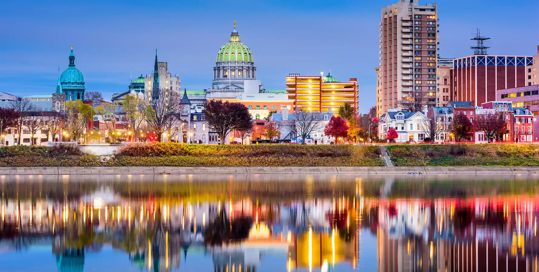 Harrisburg skyline at dusk, featuring the state capitol dome, colorful city buildings, and their reflections on the Susquehanna River.