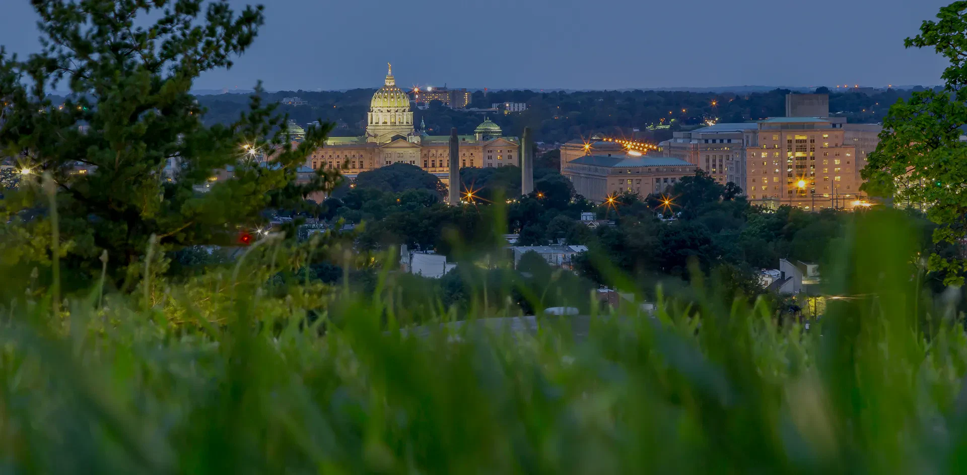 view of a city skyline at dusk, featuring a lit state capitol dome amidst greenery in the foreground and a softly glowing skyline.